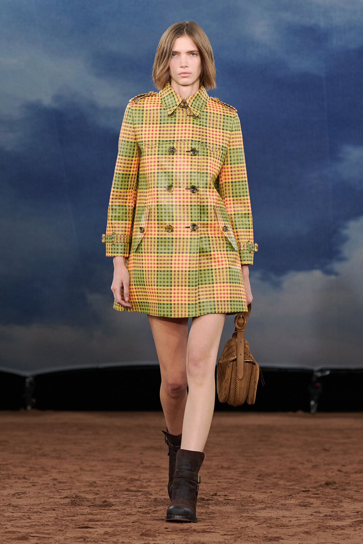 Model walking a fashion runway wearing a yellow-green plaid double-breasted coat with epaulets, brown boots, and a tan bag.
