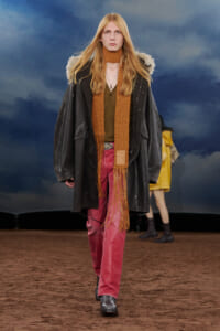 Model walking a runway in an oversized black coat with a tan fur-like collar, orange knit scarf, olive shirt, red pants, and black boots.