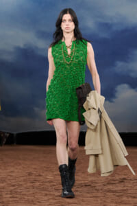 Female model walking a runway in a bright emerald-green textured sleeveless dress, black lace-up boots, and a beige trench coat draped over her arm.