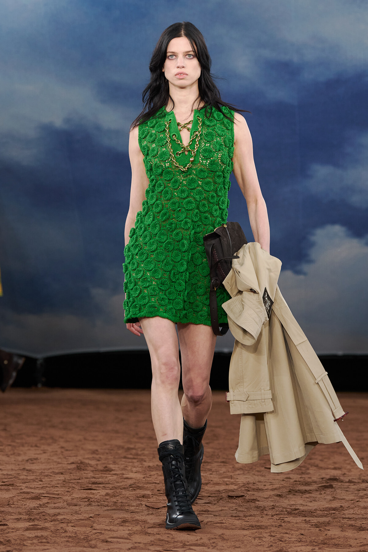 Female model walking a runway in a bright emerald-green textured sleeveless dress, black lace-up boots, and a beige trench coat draped over her arm.