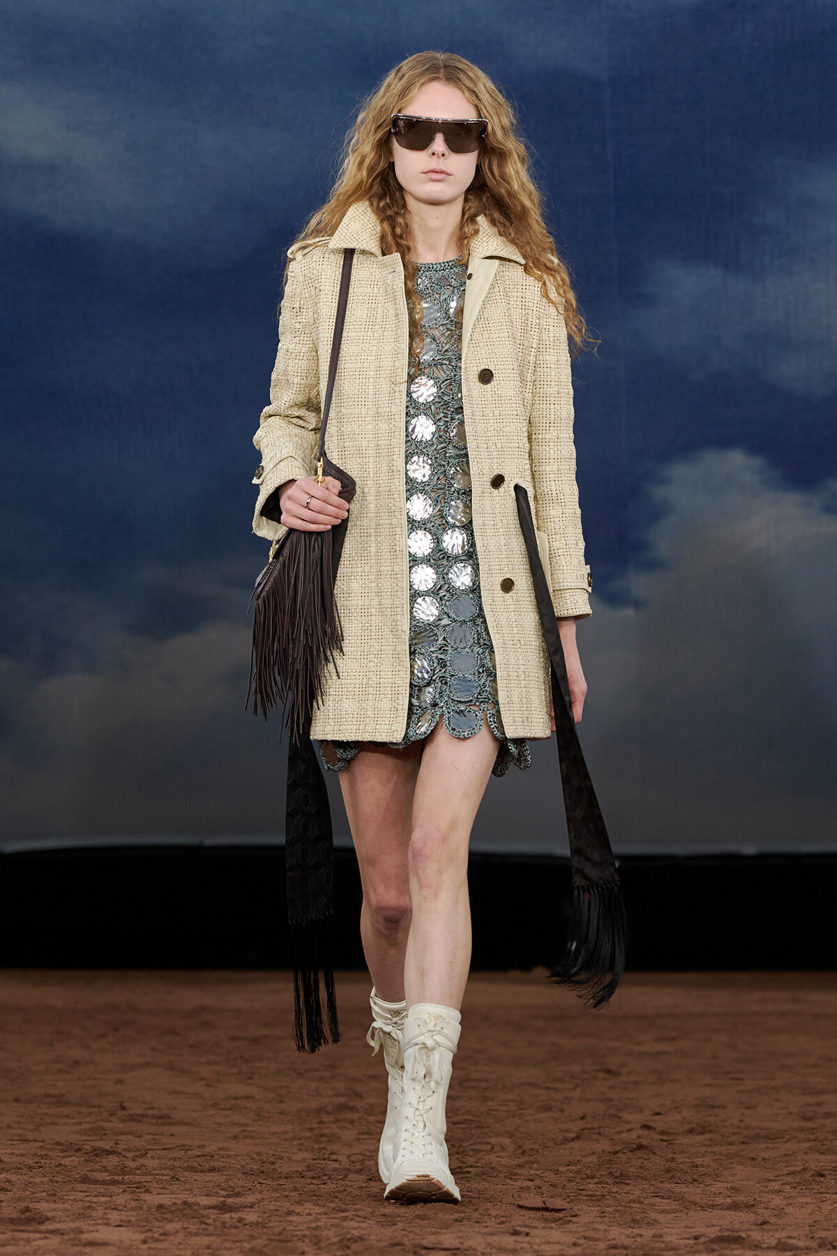 Model walking a runway in a beige textured coat over a metallic silver dress, with a fringed brown bag and oversized sunglasses.
