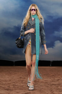 Model walking a runway in a blue denim trench, turquoise scarf, floral dress peeking underneath, and white-and-black striped sandals with a black fringed bag.