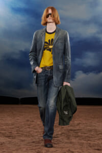 Male model walking a fashion runway in a blue denim blazer and jeans, mustard-yellow graphic tee, brown belt, red hair with sunglasses, carrying a green jacket on the side.
