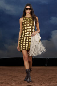 Model walking a runway in a gold circular-patterned sleeveless dress, large sunglasses, a white fringed handbag, and black lace-up boots on a dirt catwalk.