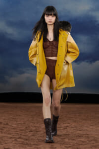 Model strides down a runway wearing a mustard-yellow oversized coat over a brown crochet top and shorts, with black lace-up boots.