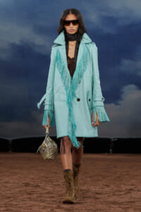 Model on a runway wearing a turquoise fringe coat with oversized sunglasses, carrying a studded handbag and wearing tan lace-up boots.