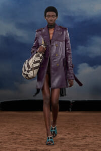Model walking the runway in a purple leather trench coat, holding a black-and-white patterned bag.