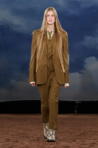Model walking a fashion runway in a brown pinstripe suit (jacket, vest, trousers) with a striped shirt and tie; snakeskin boots visible against a cloudy blue backdrop.
