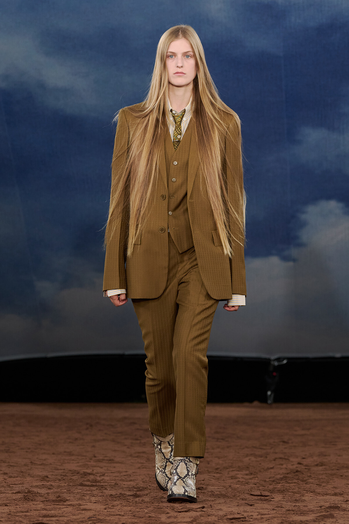 Model walking a fashion runway in a brown pinstripe suit (jacket, vest, trousers) with a striped shirt and tie; snakeskin boots visible against a cloudy blue backdrop.