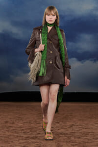 Model walking a fashion show in a brown leather coat with a green patterned scarf, cream fringe bag, and colorful sandals on a desert-like runway.