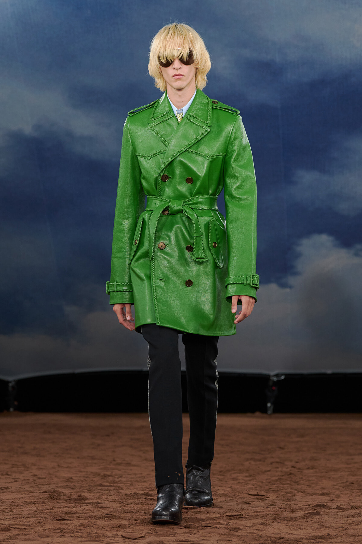 Model on runway wearing a vibrant green belted trench coat with oversized lapels and dark pants, sunglasses, against a cloudy backdrop.