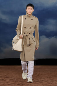Model walking a fashion runway in a beige trench coat with belt, white pants, white sneakers, and a large white shoulder bag against a cloudy blue backdrop.