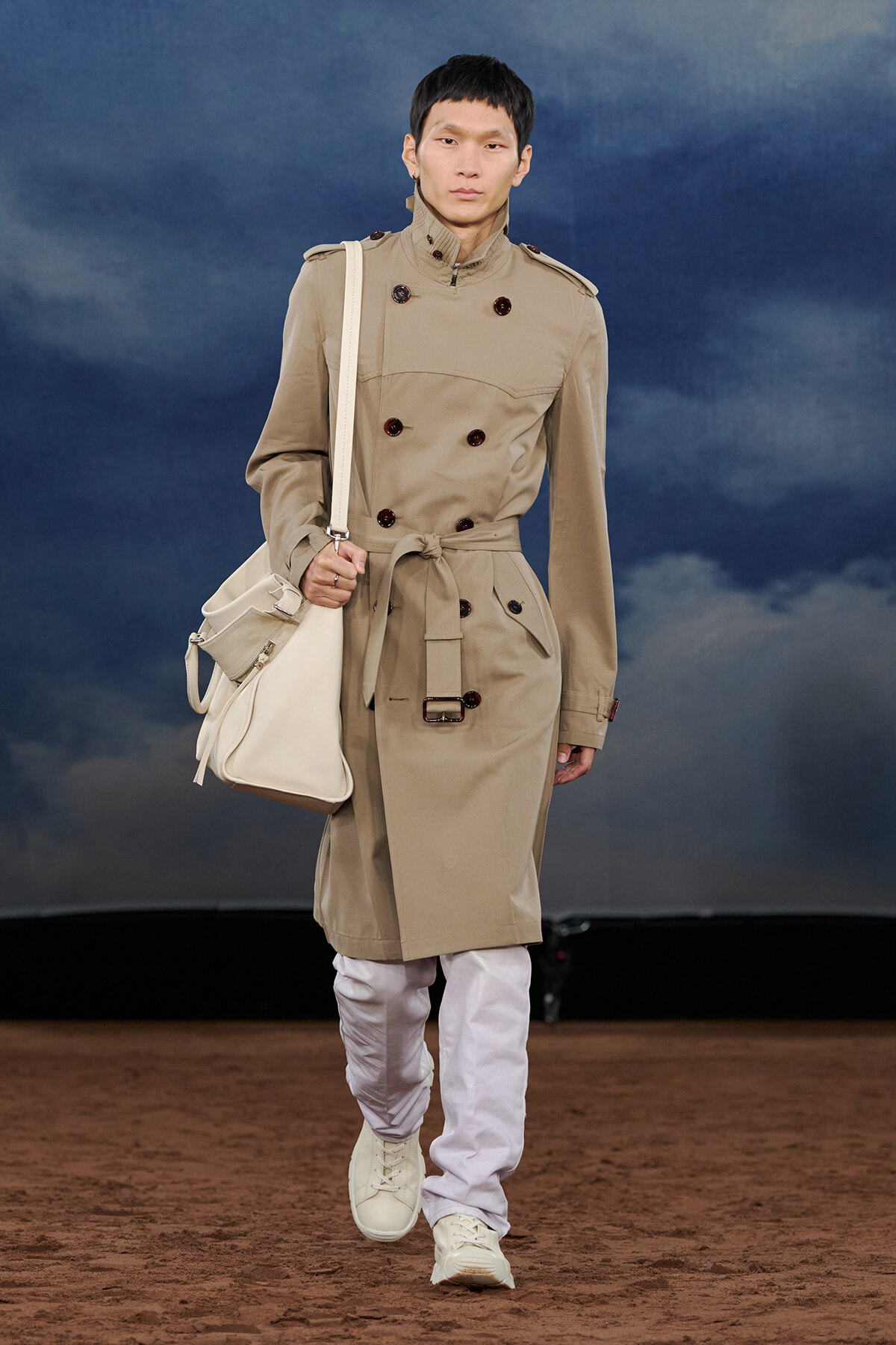 Model walking a fashion runway in a beige trench coat with belt, white pants, white sneakers, and a large white shoulder bag against a cloudy blue backdrop.