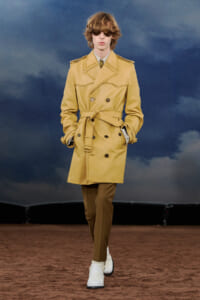 Male model walks the runway in a tan belted trench coat, brown trousers, and white boots, with dark sunglasses.