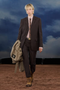 Model walking a runway in a brown pinstripe suit with striped shirt and tie, carrying a beige coat against a cloudy blue backdrop.