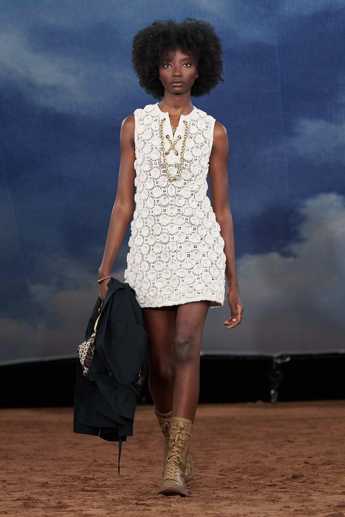 Model walks a runway in a cream crochet mini dress, layered with gold chains, tan lace-up boots, and a black jacket draped over her arm.