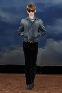Male fashion model walking on a runway in a denim bomber jacket, black pants, and sunglasses.