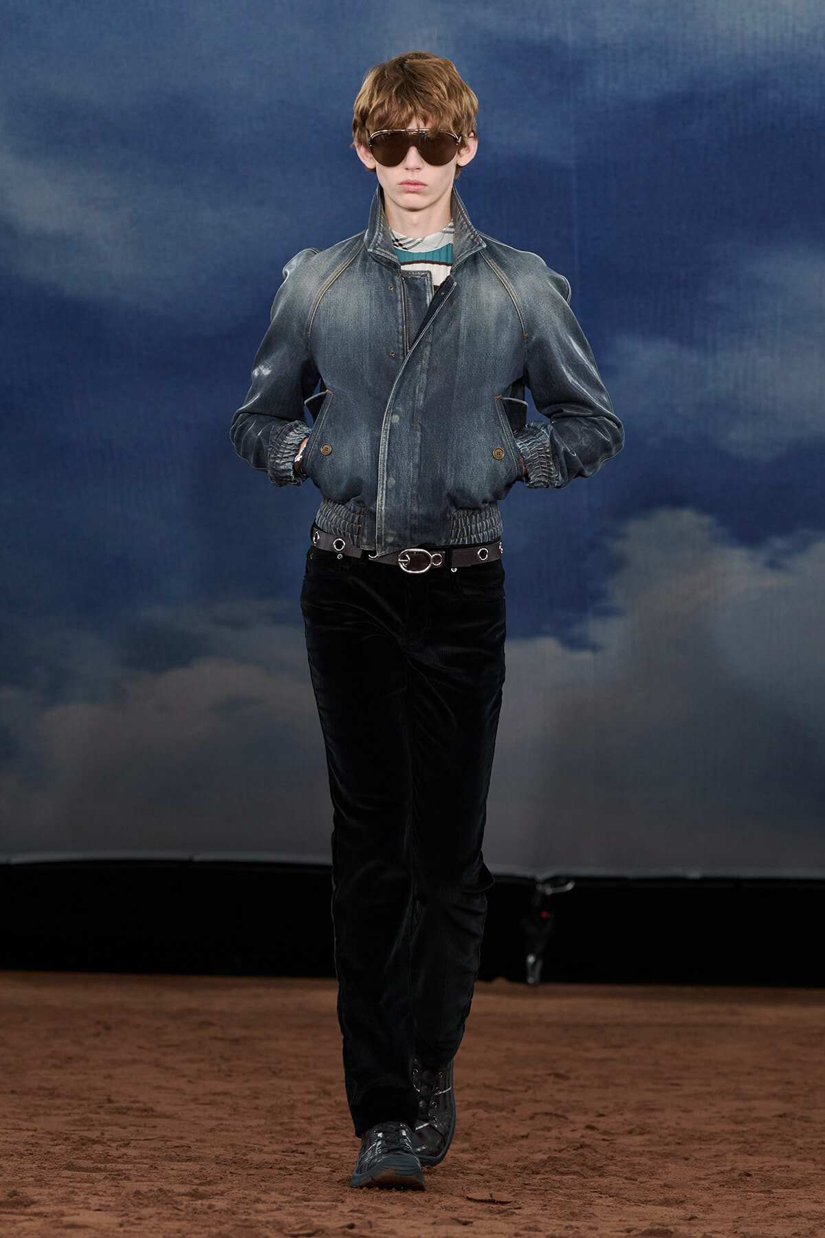 Male fashion model walking on a runway in a denim bomber jacket, black pants, and sunglasses.