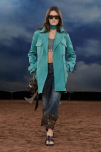 Model walking runway in turquoise coat, cropped top, baggy jeans with circular embellishments, and oversized sunglasses.