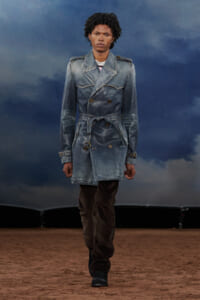 Male fashion model walking a runway in a washed denim trench coat, belted at the waist, with brown pants and black boots against a blue cloudy backdrop.