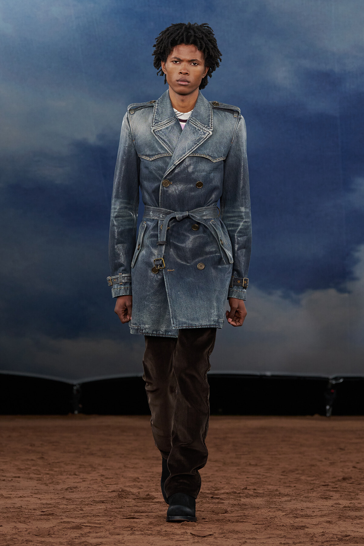 Male fashion model walking a runway in a washed denim trench coat, belted at the waist, with brown pants and black boots against a blue cloudy backdrop.