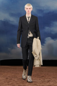 Male model walking a runway in a black suit with beige shirt and patterned tie, carrying a beige trench coat; wearing snakeskin boots.