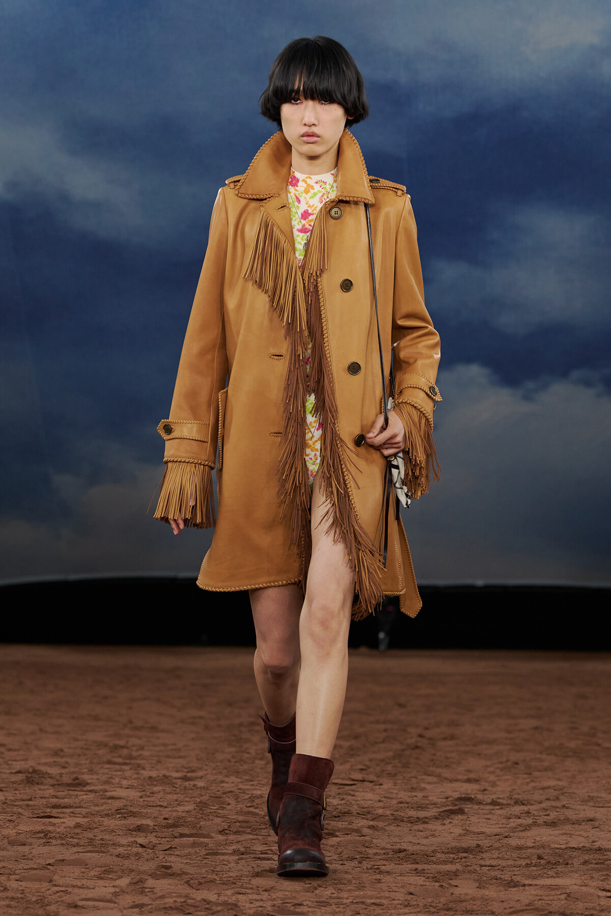 Model walking runway in a tan fringe suede coat over a floral dress, brown ankle boots, against a desert-like backdrop.