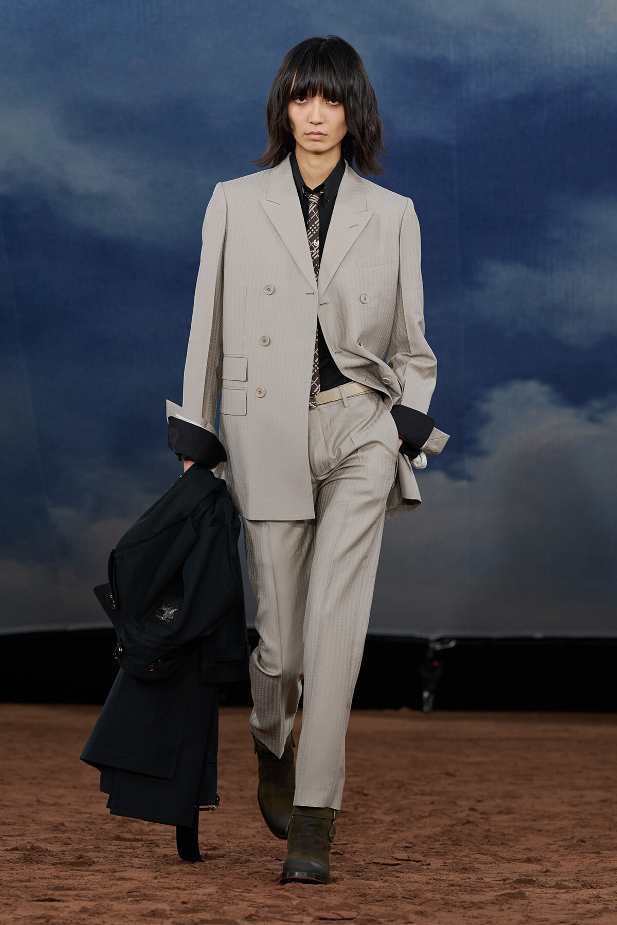 Model walking down a fashion runway in a light beige pinstripe suit, black shirt and patterned tie, holding a dark coat in one hand against a blue cloudy backdrop.