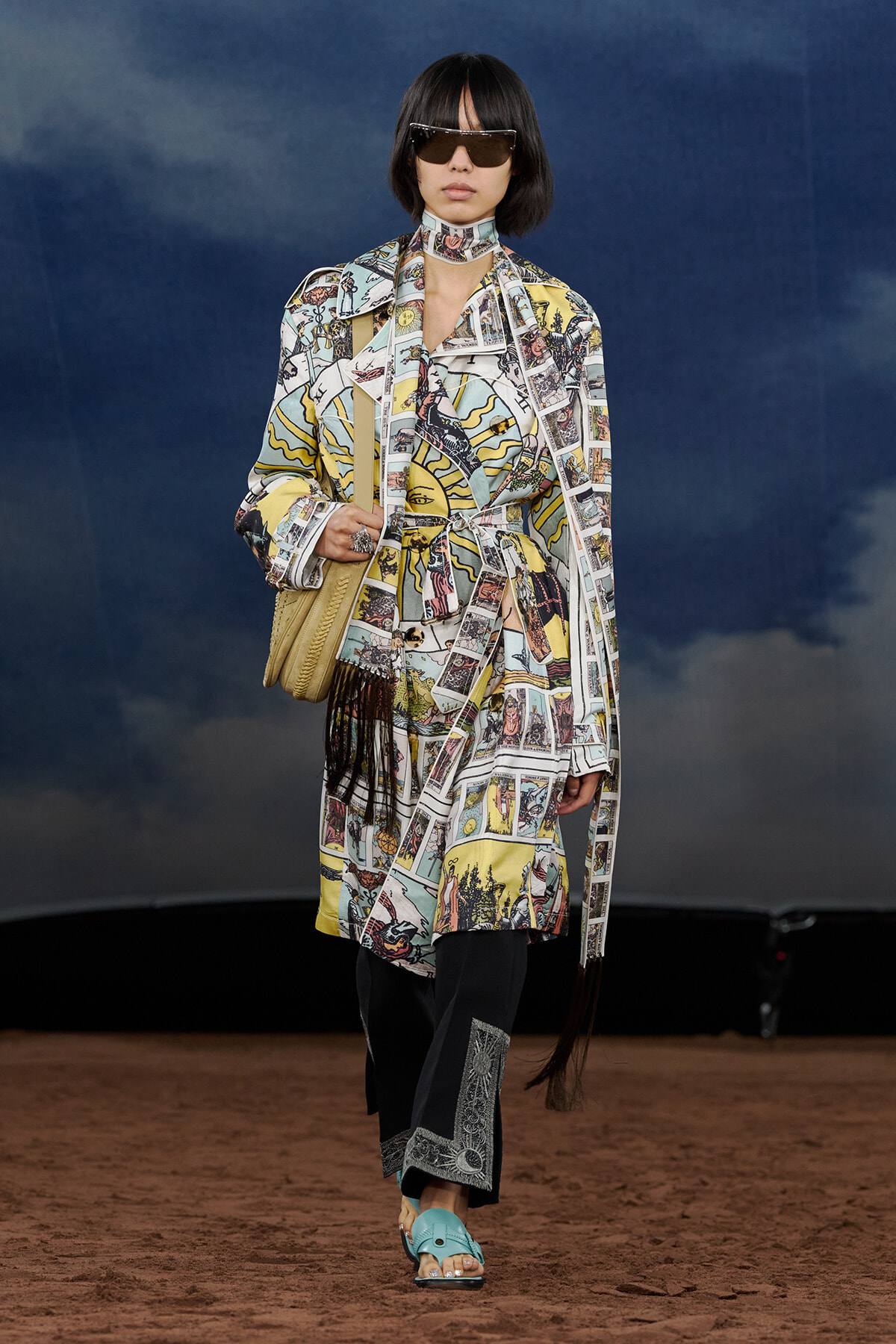 Model walking a runway in a colorful comic-strip print trench and matching dress with belt, black pants, and turquoise sandals, carrying a woven bag and wearing oversized sunglasses.