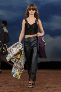 Model walking the runway in a black crochet crop top, leather pants, and a purple fringe bag, holding a printed comic-style jacket.