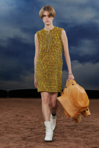 Model walks the runway in a textured yellow-green knit shift dress with a long gold chain necklace, carrying an orange jacket and wearing white boots.