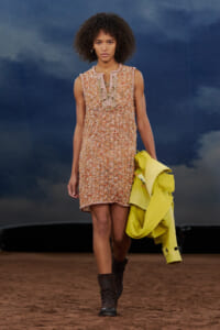 Model walks the runway in a brown textured sleeveless mini dress with a gathered neckline, holding a neon yellow jacket and wearing brown boots.