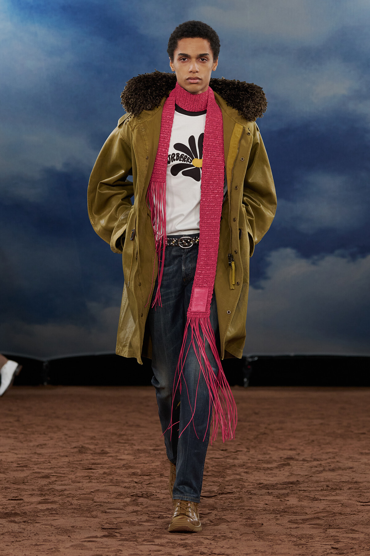 Male model walks a runway in a tan parka with shearling collar, a bright pink scarf, white graphic tee, and dark jeans.