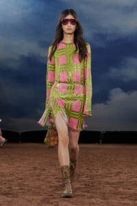 Model walking a runway in a pink-and-lime plaid sequin dress with fringe trim, pink sunglasses, and brown lace-up boots.