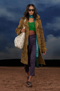 Model walking runway in a brown lace trench, green crochet crop top, and purple pants, carrying a white lace handbag and wearing oversized sunglasses