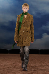 Model walking a runway in a tan paisley trench coat with belt, green scarf, and gray trousers against a blue backdrop