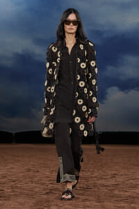 Model walking on a runway wearing a long black coat covered in circular crochet patches, black outfit, and oversized sunglasses against a cloudy blue backdrop.