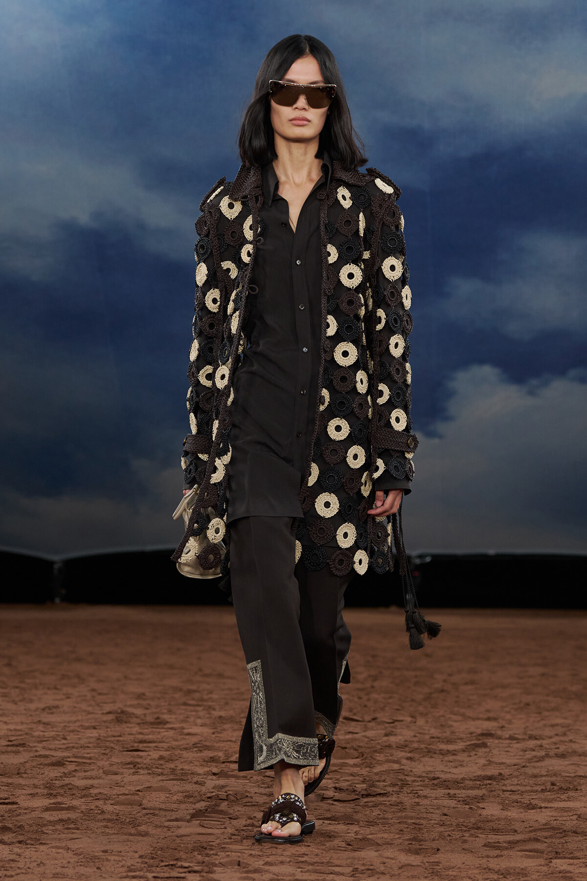 Model walking on a runway wearing a long black coat covered in circular crochet patches, black outfit, and oversized sunglasses against a cloudy blue backdrop.