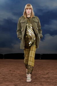 Runway model in olive utility jacket over a gold lattice dress and mustard plaid pants with circular fringe at the cuffs, walking on a brown runway.