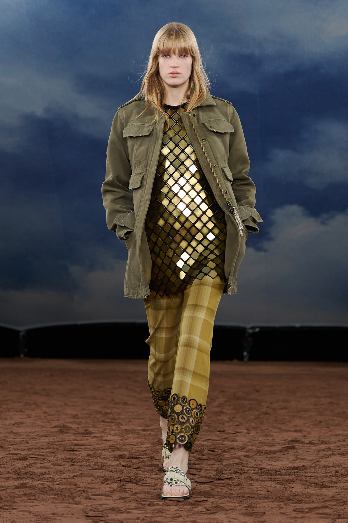 Runway model in olive utility jacket over a gold lattice dress and mustard plaid pants with circular fringe at the cuffs, walking on a brown runway.
