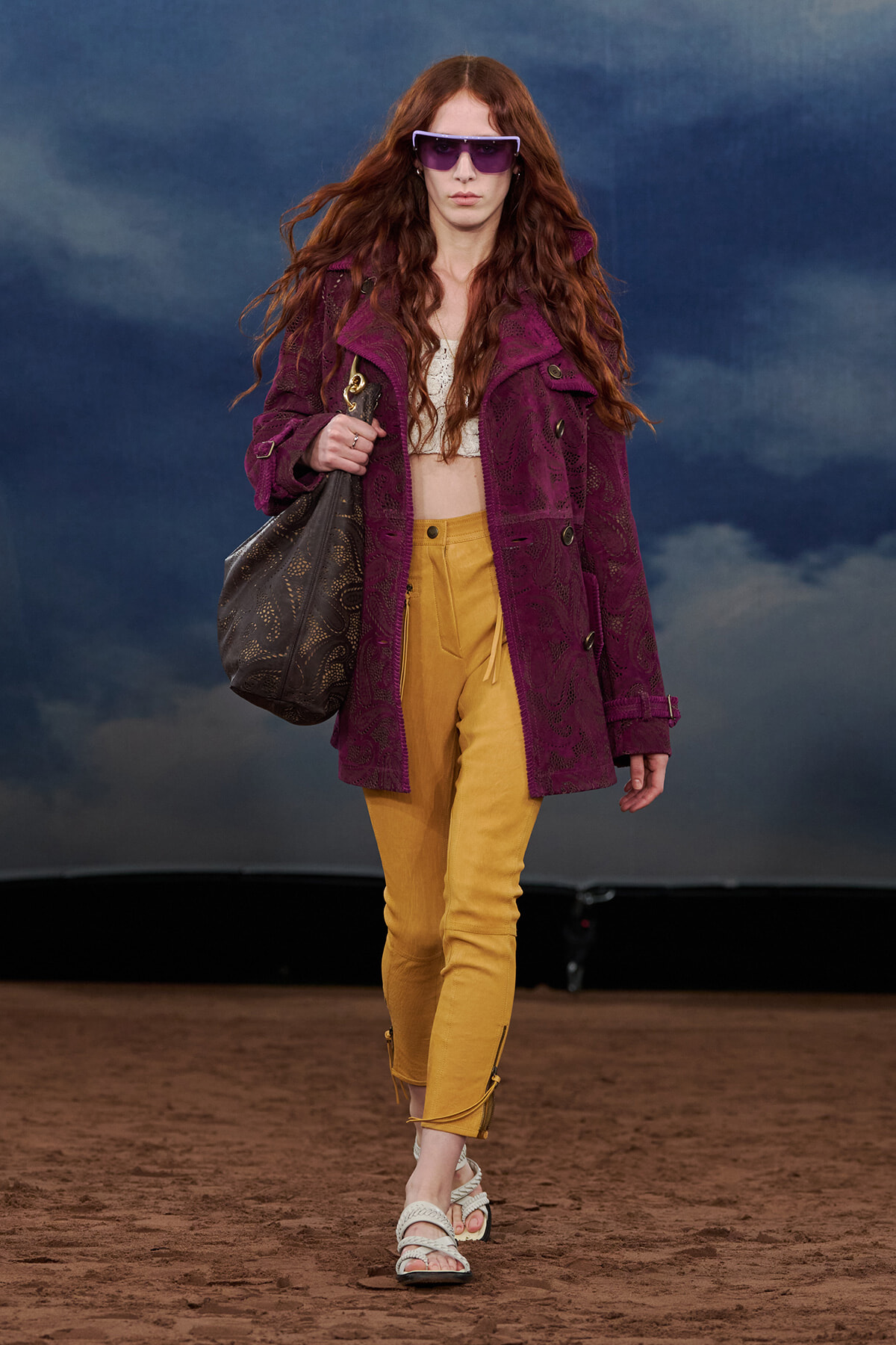 Runway model wearing a purple lace coat over a white top and mustard pants, with oversized purple sunglasses and a large black patterned bag.