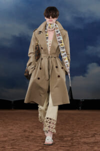 Model walking a runway in a tan belted trench coat with a comic-strip scarf and lace-trimmed pants. Sheer sunglasses complete the look.