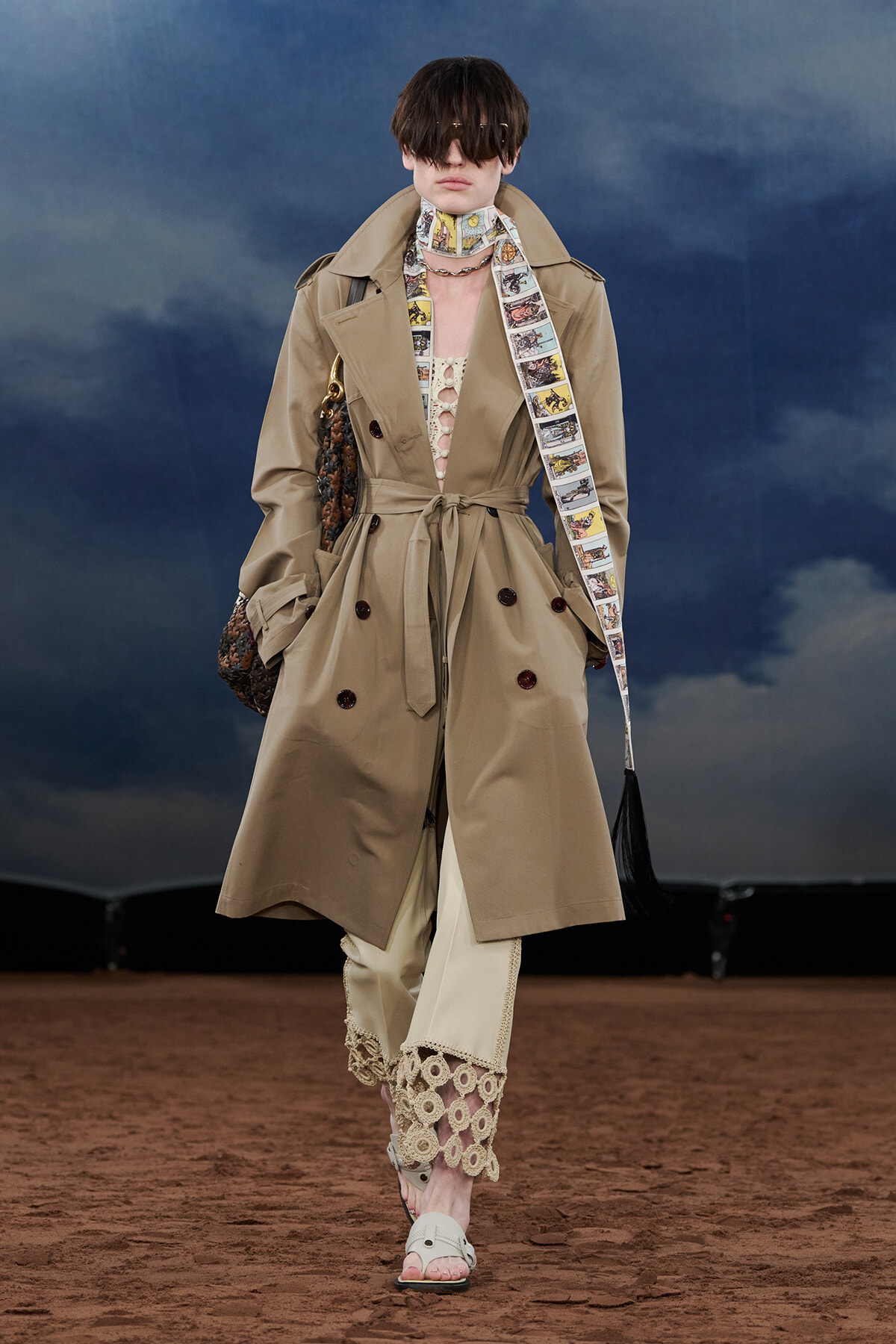 Model walking a runway in a tan belted trench coat with a comic-strip scarf and lace-trimmed pants. Sheer sunglasses complete the look.