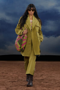 Model walking on a runway in an olive-green coat and matching pants, oversized sunglasses, black boots, and a bright pink-green crochet bag.