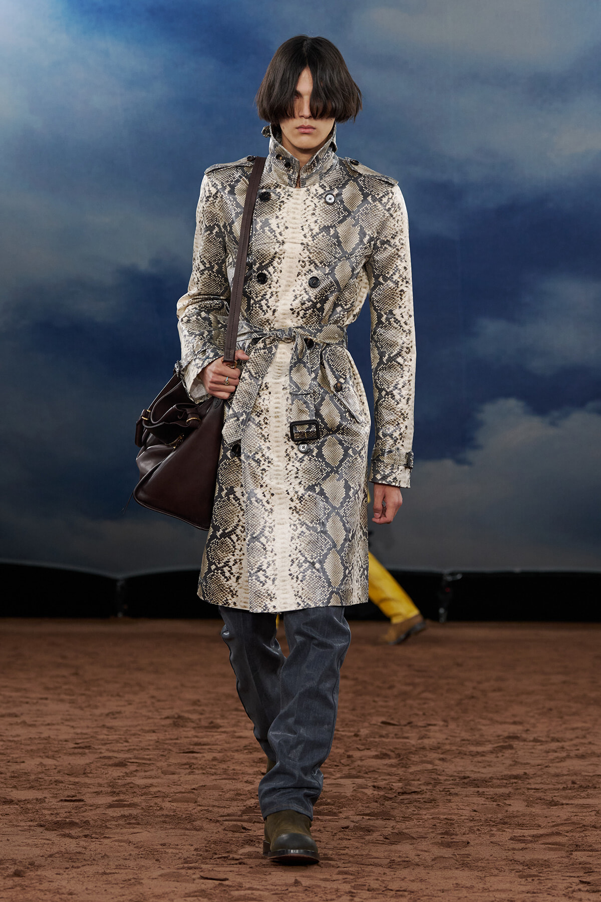 Model walks the runway wearing a snakeskin-print trench coat belted at the waist, carrying a dark brown shoulder bag.