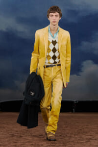 Male model walks the runway in a bright yellow suit with an argyle sweater, carrying a black coat in his left hand they style a fashion show look