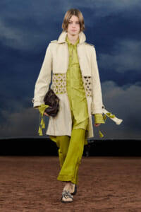 Model walking the runway in a lime-green outfit with a long cream coat featuring lace panels and tassels, carrying a dark handbag.