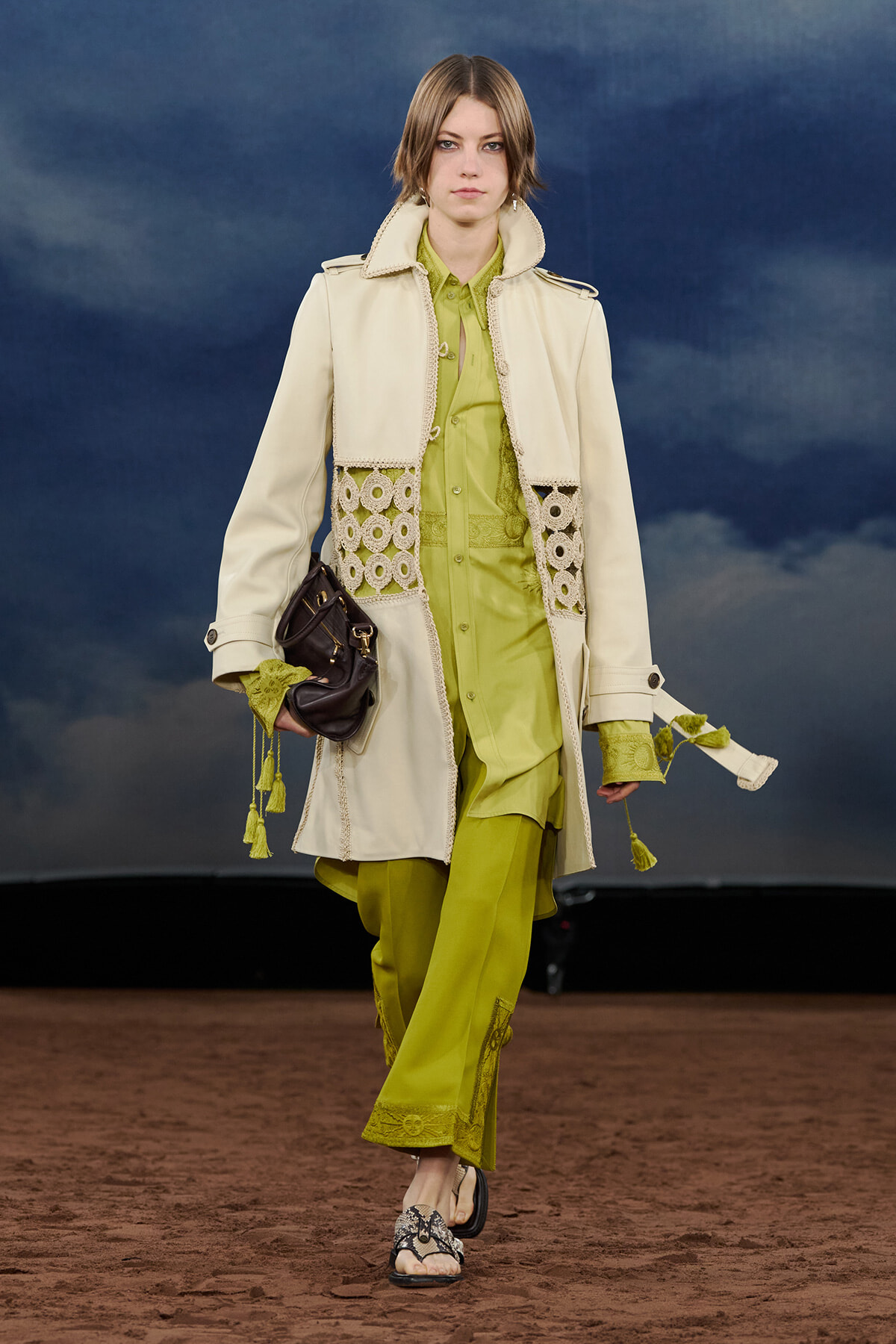 Model walking the runway in a lime-green outfit with a long cream coat featuring lace panels and tassels, carrying a dark handbag.