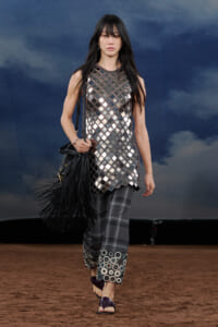 Model walking a runway in a metallic diamond-pattern tunic over plaid pants with circular cuff embellishments, carrying a black fringed handbag.