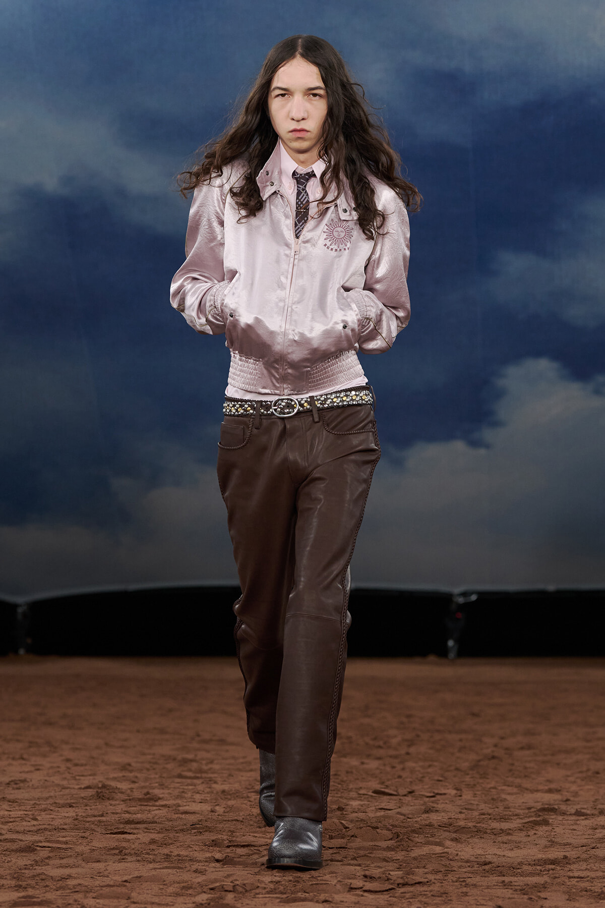Model walks a fashion runway wearing a pale pink satin bomber jacket, brown leather pants, a studded belt, and gray boots against a blue cloudy backdrop.